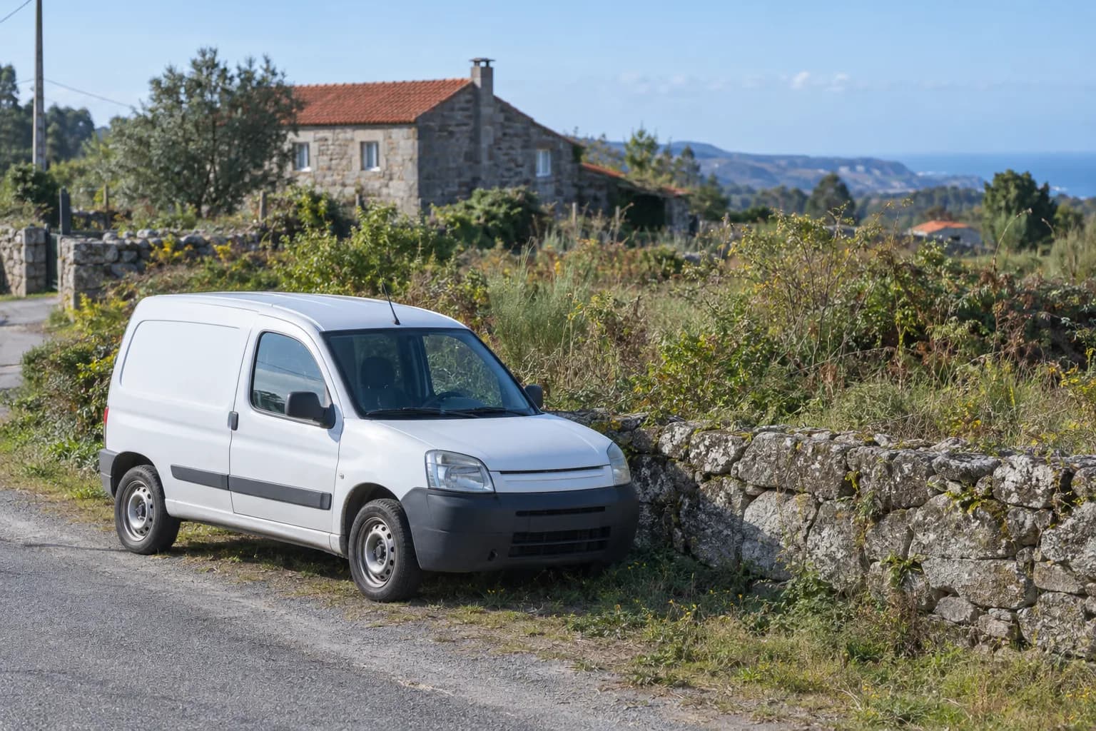 Carrinha branca, sem logótipo, estacionada junto a um muro de pedra e a um terreno com mato