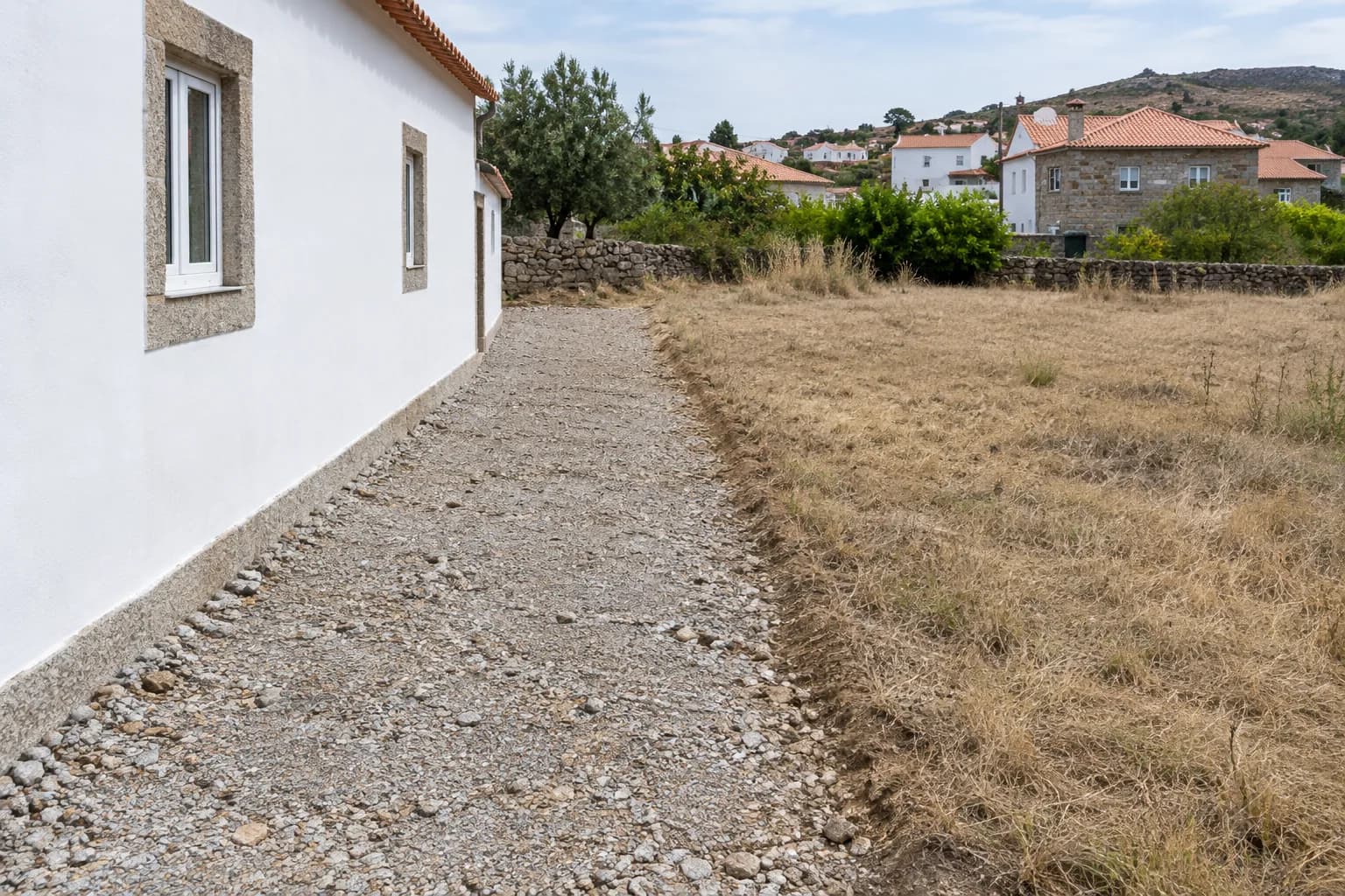 Faixa de gravilha limpa junto à parede de uma casa caiada, com erva seca ao fundo